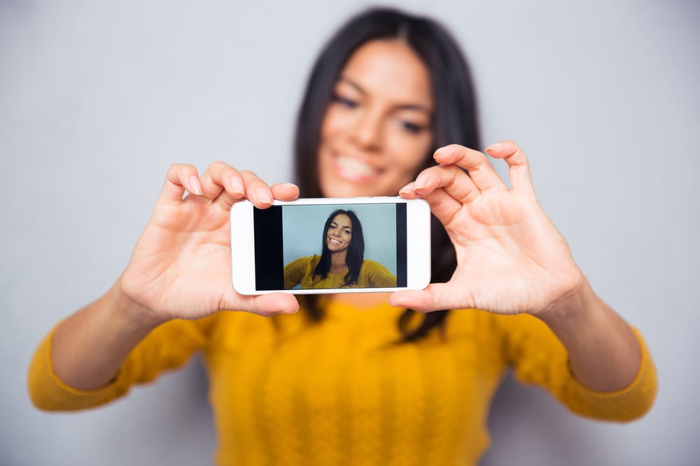 Happy woman making a selfie as a form of relatable content, using her smartphone over gray background. Focus is on smartphone.